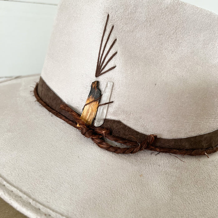Close up detail of the clear crystal, Palo Santo wood and brown strap on the Peace Hat. Made of pale taupe suede.